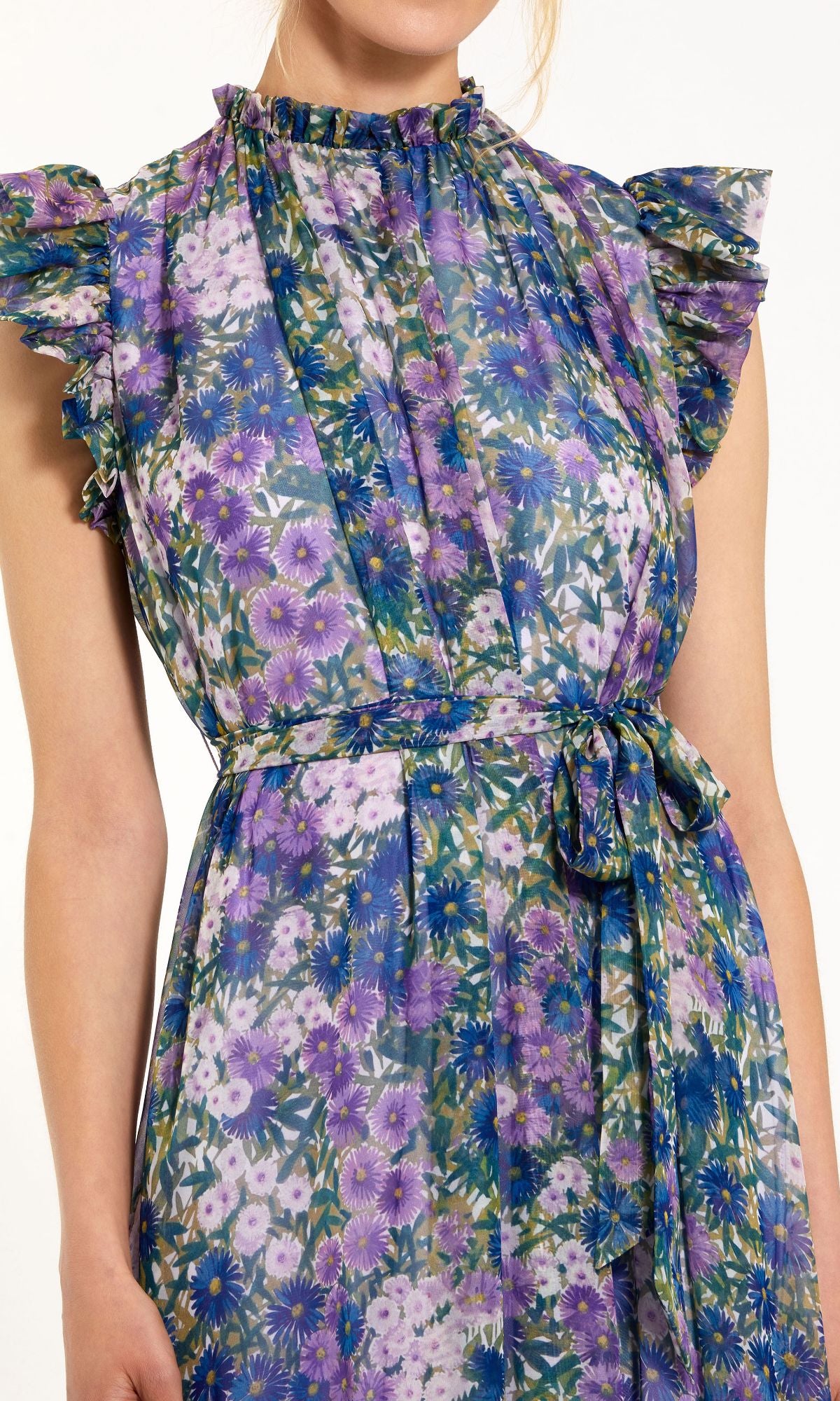 A woman wears the Lilac Floral-Print Formal Gown by Mac Duggal 56116, featuring a sleeveless, ruffled design and a matching tie belt at the waist in purple, blue, and green florals.